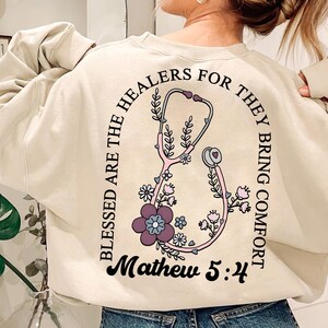 May include: Cream-colored sweatshirt with a floral stethoscope design. The design includes the text "Blessed are the healers for they bring comfort" and "Mathew 5:4". The stethoscope is pink and purple.