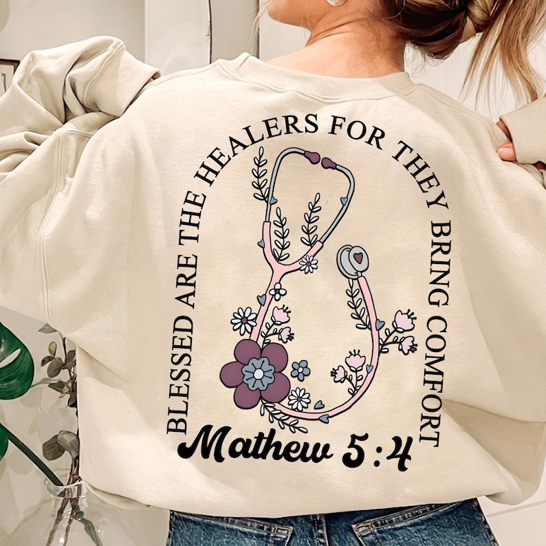 May include: Cream-colored sweatshirt with a floral stethoscope design. The design includes the text "Blessed are the healers for they bring comfort" and "Mathew 5:4". The stethoscope is pink and purple.