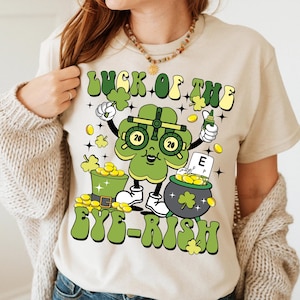 May include: A beige t-shirt with a green shamrock cartoon character wearing eye glasses and holding a pot of gold. The text "Luck of the Eye-rish" is printed on the shirt.