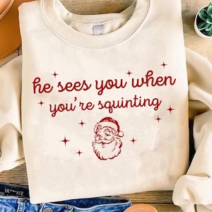 May include: A cream-coloured sweatshirt featuring the red text "he sees you when you're squinting". Below the text is a red Santa Claus graphic, with red star accents. This is a festive holiday-themed garment.