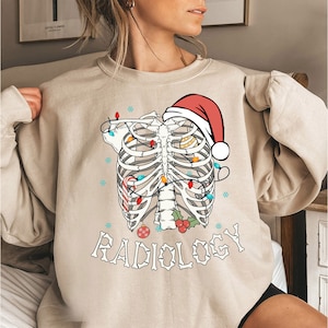 May include: A beige sweatshirt with a skeleton wearing a Santa hat and Christmas lights. The text "RADIOLOGY" is printed below the skeleton.