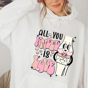 Puede incluir: Sudadera blanca con un dise&ntilde;o gr&aacute;fico rosa y negro que dice "All You Kneеd Is Love" con una ilustraci&oacute;n de rodilla de dibujos animados.