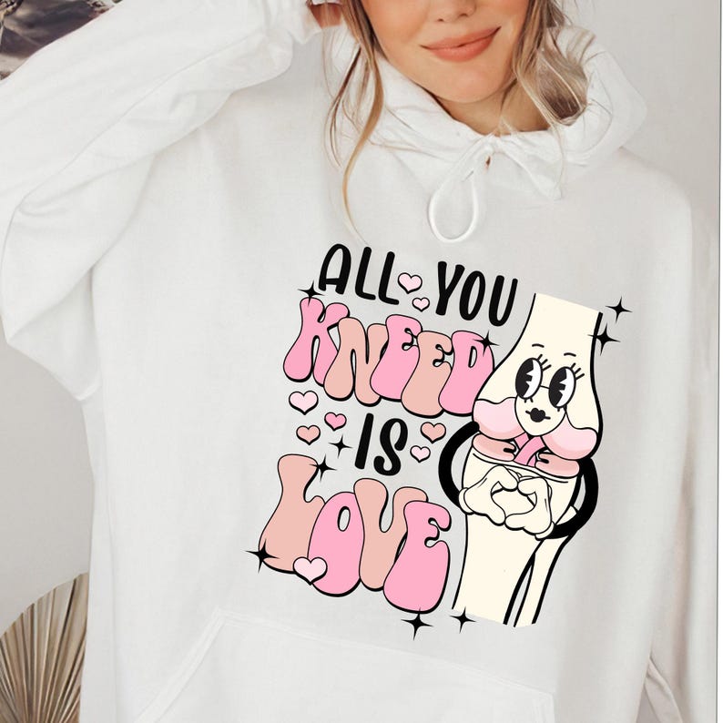 Puede incluir: Sudadera blanca con un dise&ntilde;o gr&aacute;fico rosa y negro que dice "All You Kneеd Is Love" con una ilustraci&oacute;n de rodilla de dibujos animados.