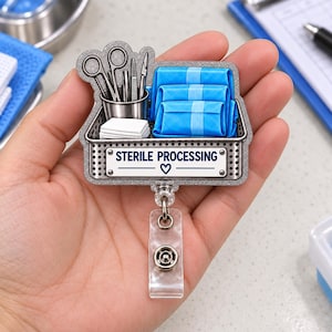 May include: A badge reel featuring a metallic basket filled with surgical instruments, white gauze, and blue wrapped sterile packs. A white rectangular label on the front displays the text STERILE PROCESSING with a small heart icon below it.