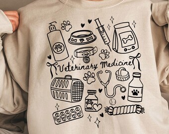 May include: A beige sweatshirt with a black graphic design featuring various veterinary medicine related items, including a stethoscope, a dog lead, a pet carrier, a pill bottle, a dog bone, a dog bowl, a syringe, and a bag of dog food. The text "Veterinary Medicine" is written in a bold, black font.