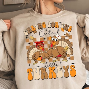 Puede incluir: Una sudadera beige con un estampado gráfico de dos pavos. El texto en la sudadera dice "Caring for the Cutest Little Turkeys".