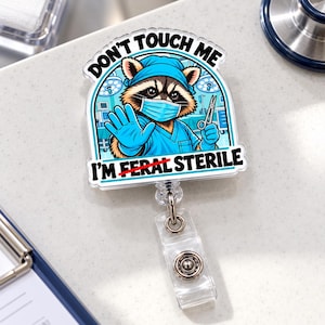 May include: A circular acrylic badge reel featuring a cartoon raccoon dressed in blue surgical scrubs, a cap, and a face mask. The raccoon holds surgical scissors and makes a stop gesture. Text reads: DON'T TOUCH ME I'M FERAL STERILE.