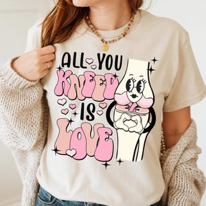 Puede incluir: Una camiseta beige con un gr&aacute;fico rosa de una rodilla de dibujos animados con el texto "All You Knead Is Love" en letras rosas.