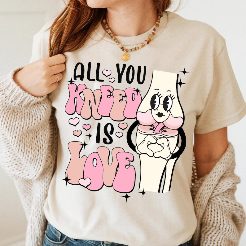 Puede incluir: Una camiseta beige con un gr&aacute;fico rosa de una rodilla de dibujos animados con el texto "All You Knead Is Love" en letras rosas.