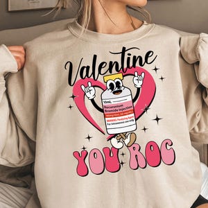 Może przedstawiać: Beżowa bluza z różowym sercem i kreskówkową butelką z napisem "Valentine You Roc" w różowych literach.