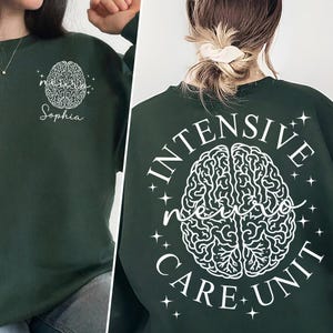 Peut inclure: Un sweat-shirt vert foncé avec un motif de cerveau blanc et le texte "INTENSIVE NEURO CARE UNIT" au dos. Le devant du sweat-shirt présente un motif de cerveau plus petit et le texte "Sophia".