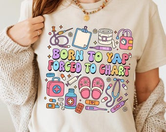 May include: Beige t-shirt with a colourful graphic featuring medical tools and the text "BORN TO YAP FORCED TO CHART". The design includes a stethoscope, syringe, pills, and a first aid kit.