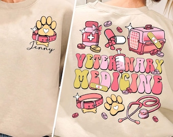 May include: A light beige sweatshirt featuring a graphic design of veterinary medicine themed illustrations. The design includes a paw print, a collar, a pill bottle, a syringe, a stethoscope, a pet carrier, and the text "Veterinary Medicine". The sweatshirt also has the name "Jenny" printed on the front.