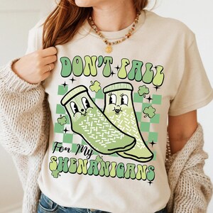 以下が含まれることがあります： 薄いベージュのTシャツに、緑と白の漫画の靴下2つが顔を描いたグラフィックがプリントされています。"Don't Fall For My Shenanigans"というテキストが緑と白の文字でプリントされています。