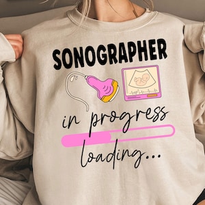 Puede incluir: Sudadera beige con la palabra "SONOGRAPHER" en negro. Debajo, se lee "in progress loading..." en una fuente de escritura. El diseño incluye una sonda de ultrasonido rosa y una imagen de ultrasonido rosa y blanca.