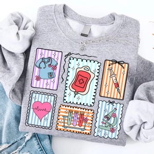 May include: A grey sweatshirt with colourful medical-themed patches. Patches include a nurse's uniform, a blood bag, a syringe, a heart monitor, test tubes, and a microscope. The patches have striped backgrounds and decorative borders. A silver necklace is draped on the sweatshirt.