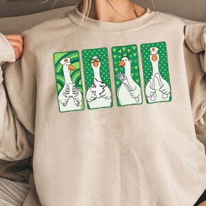 Op de afbeelding: Beige sweatshirt met vier cartoon ganzen in groene kaders. Elke gans wordt afgebeeld in een andere medische rol, met een verpleegsterspet en een ander die een skelet vasthoudt. Het ontwerp heeft een grillige en humoristische stijl.