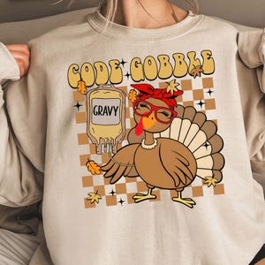 Può includere: Felpa beige con grafica a tema Ringraziamento. Il design presenta un tacchino dei cartoni animati con occhiali e un fiocco rosso, con le parole "CODE GOBBLE" sopra. È inclusa anche una sacca di salsa.