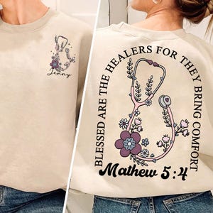 May include: Beige sweatshirt with a stethoscope and floral design. The front features a small stethoscope graphic with the name "Jenny". The back has a larger stethoscope design with the text "Blessed are the healers for they bring comfort" and "Mathew 5:4".