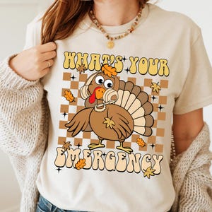 May include: Beige t-shirt with a cartoon turkey graphic. The turkey has a headset and is set against a brown and tan checkered background. The text "WHAT'S YOUR EMERGENCY" is above and below the turkey.
