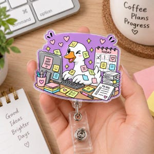 May include: A clear acrylic badge reel featuring a cartoon duck at a desk with a computer, a calendar, and sticky notes. The computer screen displays the text 'YOU GOT THIS!' and the duck is surrounded by pink hearts and yellow stars on a purple background.