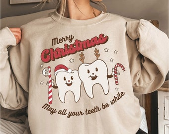 Op de afbeelding: Een beige sweatshirt met een kerstthema met daarop twee cartoon-tanden die een kerstmanmuts en rendiergewei dragen. De tekst "Merry Christmas" en "May all your teeth be white" staat op het sweatshirt gedrukt.