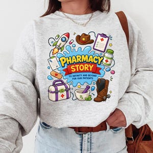 May include: A light gray crewneck sweatshirt featuring a colorful graphic with the text PHARMACY STORY TO INFINITY AND BEYOND FOR OUR PATIENTS. The design includes a rocket, cowboy hat, clipboard, pill bottle, bandage, medical kit, and boot.