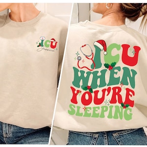 Può includere: Una felpa beige con un design a tema natalizio. La parte posteriore della felpa dice "ICU when you're sleeping" con uno stetoscopio, un cappello di Babbo Natale e agrifoglio. La parte anteriore della felpa presenta una versione più piccola del design con il nome "Jessica".