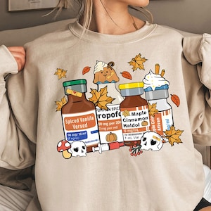 May include: A beige sweatshirt with a graphic print of four vials labeled "Spiced Vanilla Versed", "Pumpkin Spice Propolol", "Maple Cinnamon Haldol", and "Apple Crisp Ativan". The vials are surrounded by fall-themed imagery, including leaves, pumpkins, and a skull.