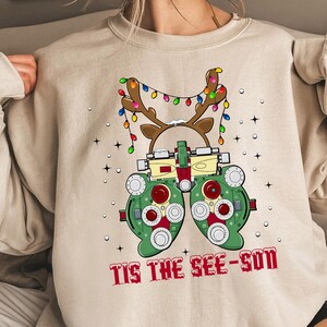 Könnte beinhalten: Beiges Sweatshirt mit weihnachtlichem Motiv. Das Design zeigt eine grüne und rote Phoropterbrille mit Rentiergeweih und einer Lichterkette. Der Text "TIS THE SEE-SON" ist in Rot gedruckt.