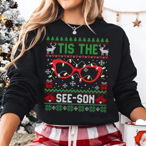 Könnte beinhalten: Schwarzer Weihnachtspullover mit grünem, rotem und weißem Muster. Der Pullover zeigt eine rote Brille mit Lichtern und dem Text "Tis the See-Son".
