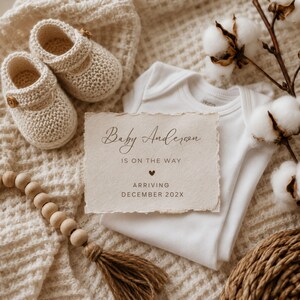 May include: A pair of cream-colored knit baby booties, a white cotton bodysuit, and a textured paper announcement card that reads Baby Anderson IS ON THE WAY with a heart icon and ARRIVING DECEMBER 202X, accompanied by wooden beads and cotton stems.
