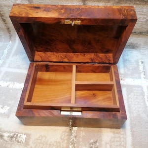 Peut inclure: Une boîte rectangulaire en bois avec un extérieur en loupe de bois poli et un couvercle à charnière. L'intérieur présente une doublure en bois clair avec un plateau amovible divisé en deux sections. Un loquet en laiton et une clé sont fixés.