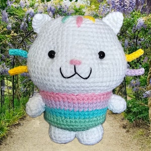 Peut inclure: Poup&eacute;e chat au crochet blanc faite &agrave; la main, avec des yeux noirs, un nez rose et un sourire cousu. La poup&eacute;e a des accents color&eacute;s sur la t&ecirc;te et un pull arc-en-ciel pastel ray&eacute;. Le chat a de petites oreilles blanches.