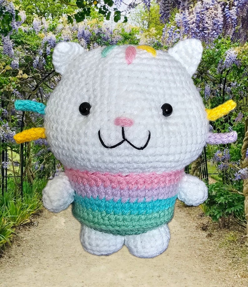 Peut inclure: Poup&eacute;e chat au crochet blanc faite &agrave; la main, avec des yeux noirs, un nez rose et un sourire cousu. La poup&eacute;e a des accents color&eacute;s sur la t&ecirc;te et un pull arc-en-ciel pastel ray&eacute;. Le chat a de petites oreilles blanches.