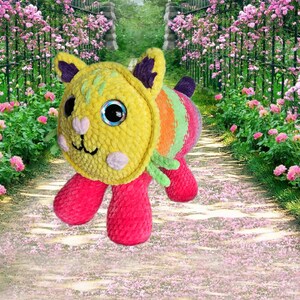 Peut inclure: Un chat en peluche avec une t&ecirc;te jaune, un corps ray&eacute; arc-en-ciel et des pattes roses. Le chat a de grands yeux bleus. L'arri&egrave;re-plan montre un sentier de jardin bord&eacute; de roses roses.