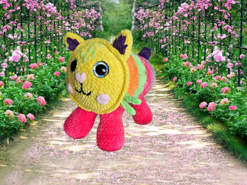 Peut inclure: Un chat en peluche avec une t&ecirc;te jaune, un corps ray&eacute; arc-en-ciel et des pattes roses. Le chat a de grands yeux bleus. L'arri&egrave;re-plan montre un sentier de jardin bord&eacute; de roses roses.
