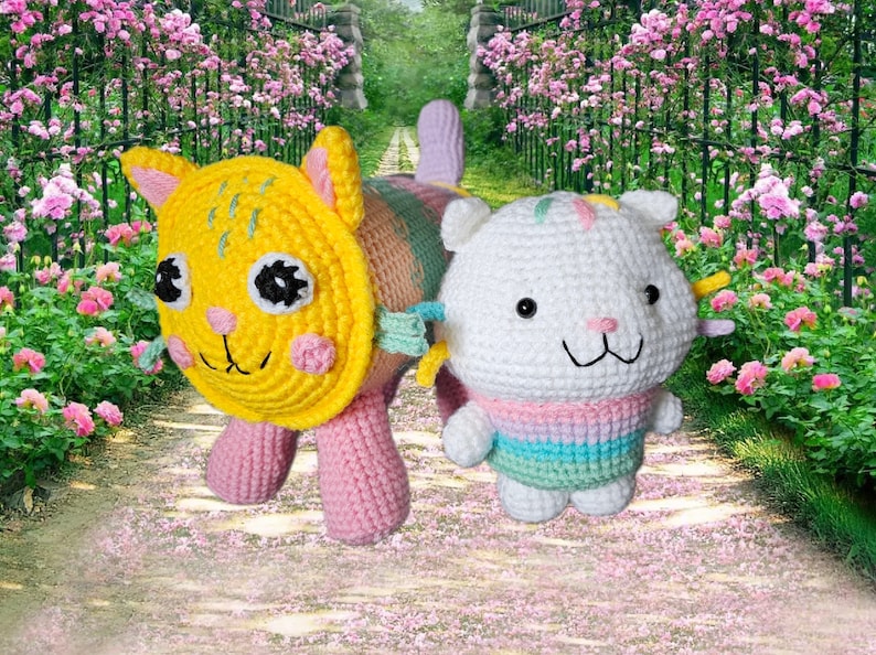 Peut inclure: Deux poup&eacute;es chats crochet&eacute;es. L'une est jaune avec des pattes roses, l'autre est blanche avec un pull &agrave; rayures pastel. Les deux ont des yeux noirs et des visages souriants. L'arri&egrave;re-plan est un chemin avec des fleurs roses et une cl&ocirc;ture en fer forg&eacute;.