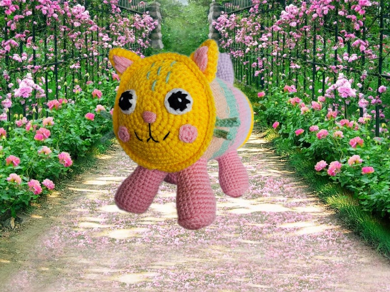 Peut inclure: Une poup&eacute;e chat au crochet faite &agrave; la main, avec une t&ecirc;te jaune, des joues roses et des yeux noirs. Le corps est ray&eacute; de couleurs pastel et les pattes sont roses. Le chat est sur un chemin avec des p&eacute;tales de fleurs roses.