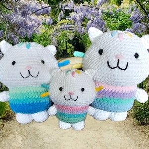 May include: Three white crochet cat dolls with colorful rainbow accents. The dolls have black eyes, pink noses, and smiling mouths. The dolls are of different sizes, with the smallest in the center. The background features a garden with purple flowers.