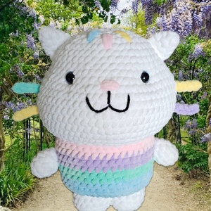 Peut inclure: Une peluche chat au crochet blanc faite &agrave; la main, avec des yeux noirs, un nez rose et une bouche souriante. Le chat a des accents color&eacute;s sur la t&ecirc;te et les oreilles, et un pull ray&eacute; arc-en-ciel. La peluche est sur un chemin avec un fond de jardin.