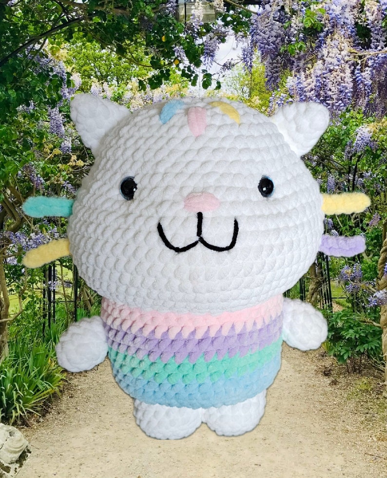 Peut inclure: Une peluche chat au crochet blanc faite &agrave; la main, avec des yeux noirs, un nez rose et une bouche souriante. Le chat a des accents color&eacute;s sur la t&ecirc;te et les oreilles, et un pull ray&eacute; arc-en-ciel. La peluche est sur un chemin avec un fond de jardin.
