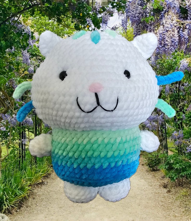 Peut inclure: Un chat en peluche fait &agrave; la main, avec une t&ecirc;te et un corps blancs, des yeux noirs, un nez rose et une bouche souriante. Le bas du corps passe du vert d'eau au bleu. Le chat a de petites oreilles et des accents bleus et verts. Le jouet est fait de fil doux et textur&eacute;.