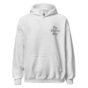 Op de afbeelding: Een lichtgrijze hoodie met een voorzak en een capuchon. De tekst "The Skyline Boys" is in zwart script gedrukt op de linkerbovenkant van de borst. De hoodie is gemaakt van een zacht, comfortabel materiaal.