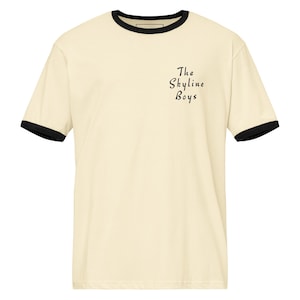 May include: A cream-coloured t-shirt with black trim around the collar and sleeves. The text "The Skyline Boys" is printed in black script on the front. The shirt has a classic, casual style.