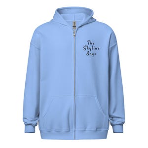 Op de afbeelding: Een lichtblauwe hoodie met ritssluiting, capuchon en voorzakken. De tekst "The Skyline Boys" is in zwart script op de borst gedrukt. De hoodie heeft een volledige ritssluiting en lange mouwen.