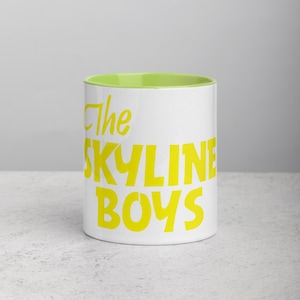 May include: White ceramic mug with a bright green interior. The mug features the text "The SKYLINE BOYS" in bold yellow letters. The mug is set against a neutral background.