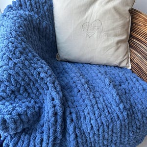 May include: A chunky knit blanket in a solid blue colour with a thick, looped texture. Resting on top is a square, off-white throw pillow featuring a centred heart-shaped line drawing and the text "Ivy Cottage" printed in a thin, dark font.