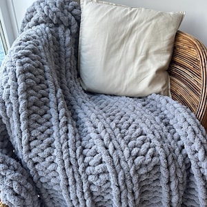 May include: A gray chunky knit throw blanket draped over a wicker chair with a beige pillow. The blanket has a cable knit pattern and is made of soft, fluffy yarn.