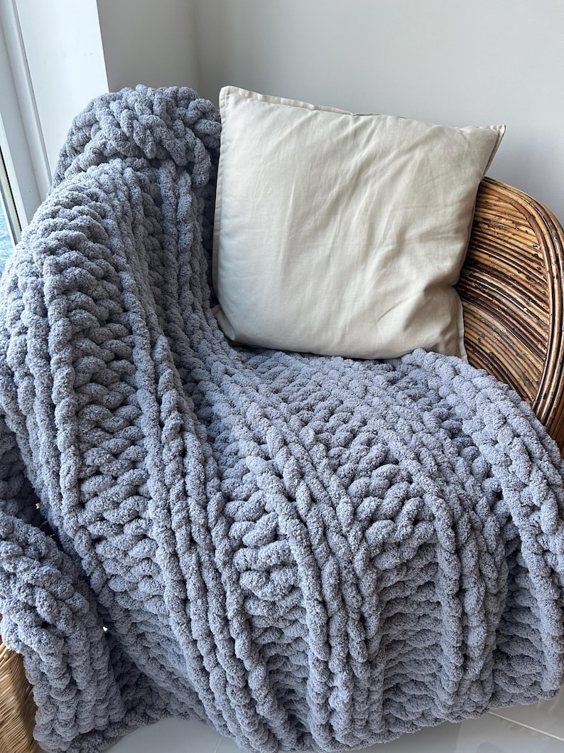 May include: A gray chunky knit throw blanket draped over a wicker chair with a beige pillow. The blanket has a cable knit pattern and is made of soft, fluffy yarn.